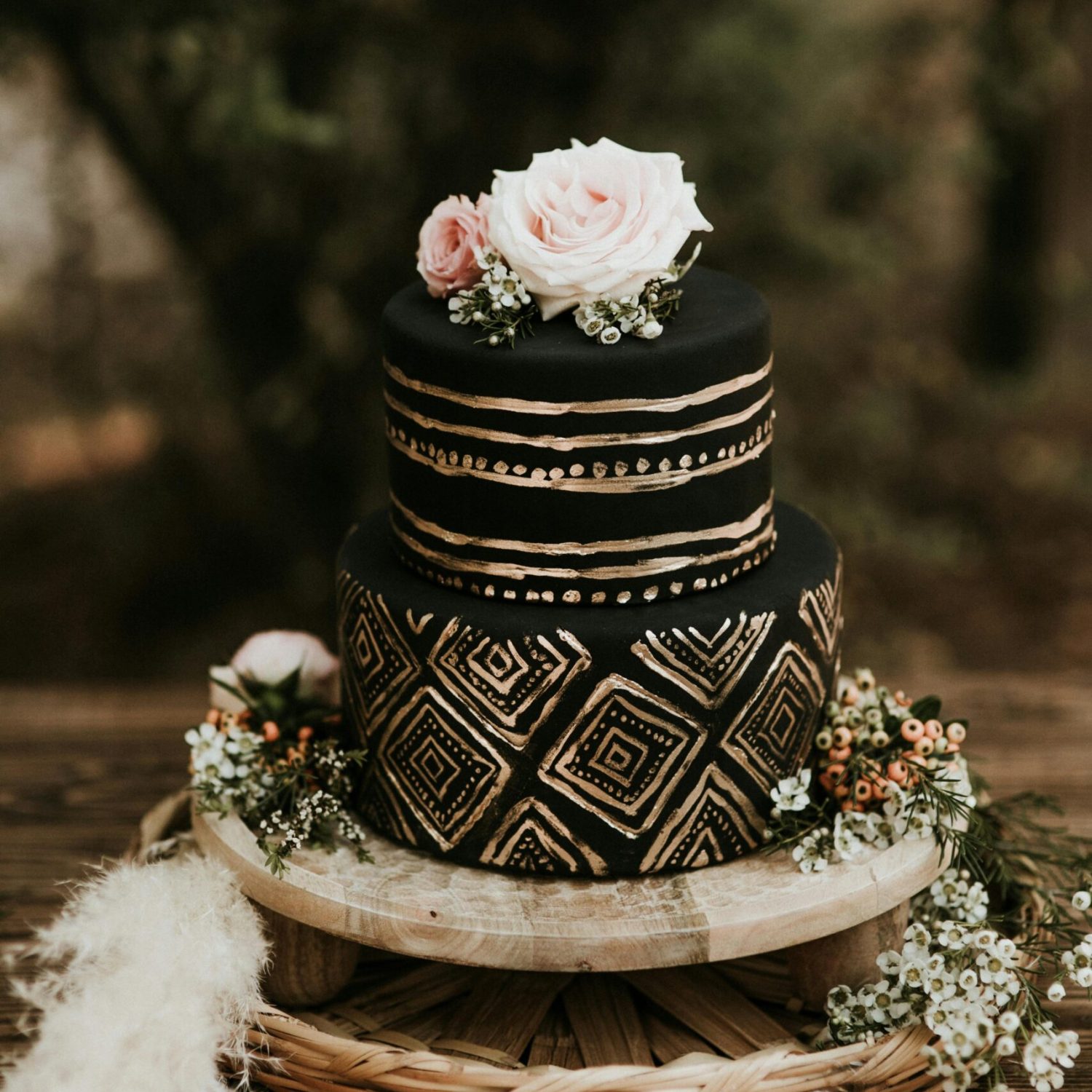 A stunning black and gold wedding cake with floral decorations on a wooden table outdoors.